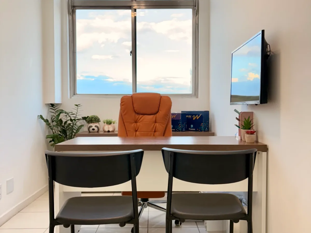 Consultório com mesa de atendimento, cadeiras e janela ampla com vista para o céu